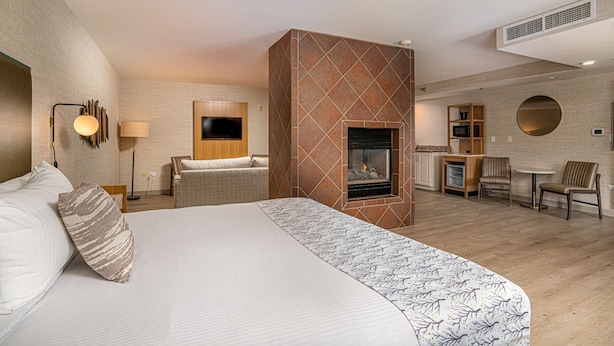 Habitación de hotel con una cama grande, chimenea, televisor montado en la pared, escritorio con sillas y una pequeña cocina.