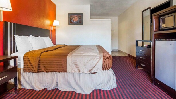 A hotel room with a bed, a desk, a dresser, a refrigerator, and a painting on the wall. - Rodeway Inn at Lake Powell (Page)