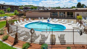 Une piscine dotée de chaises longues et d’une terrasse délimitée par une clôture, le tout entouré de bâtiments et de verdure.