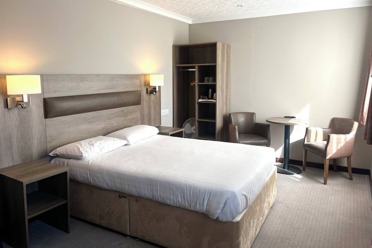 Quarto de hotel com cama, mesas de cabeceira, guarda-roupa, escrivaninha e duas cadeiras.