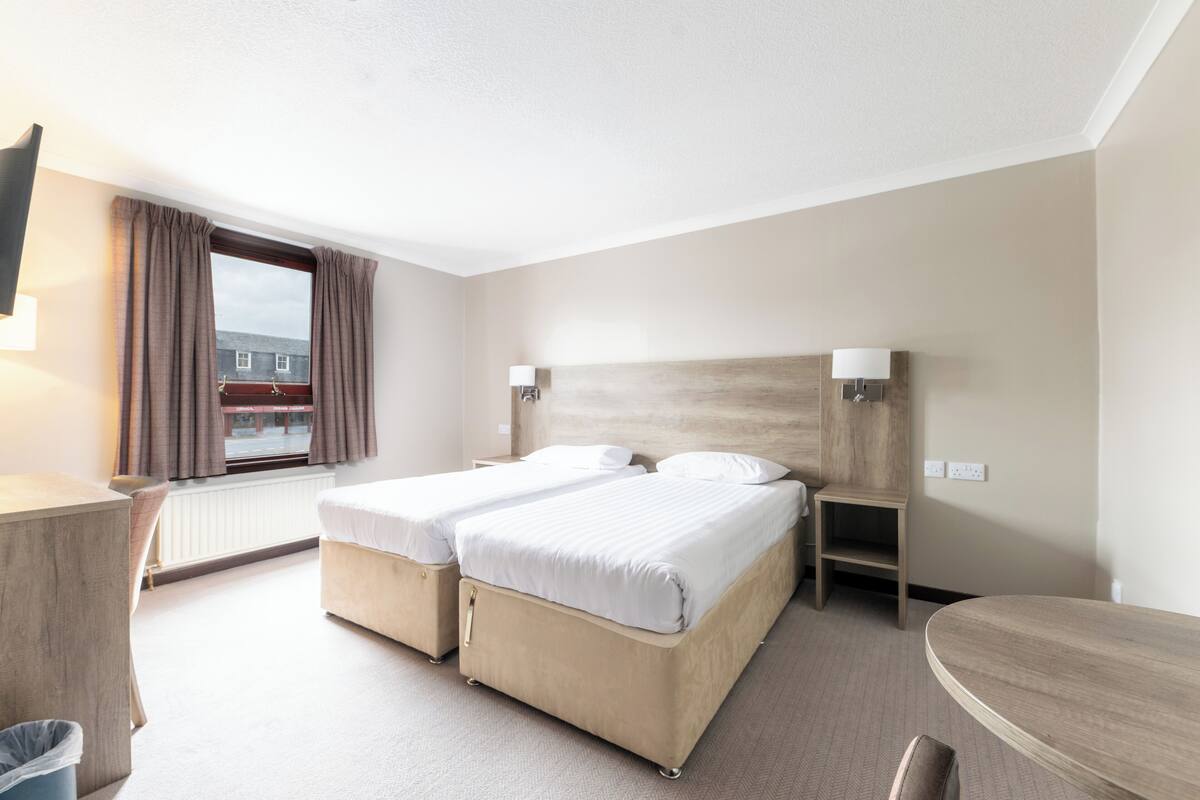 Quarto de hotel com duas camas, uma escrivaninha, uma cadeira, uma mesinha e uma janela com cortinas.