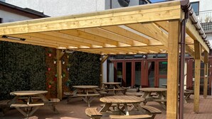 An outdoor wooden pergola with picnic tables, a brick patio, and a wall with climbing plants. - The Priory Hotel (Beauly)