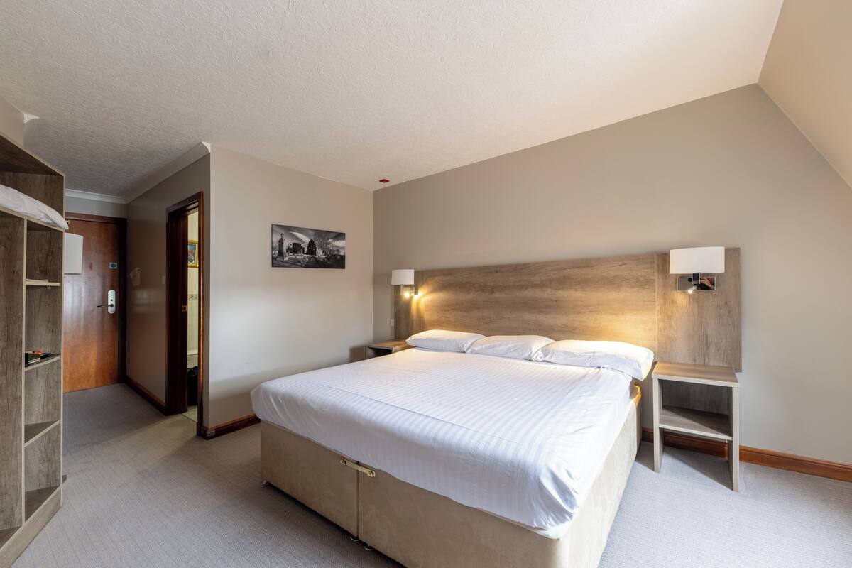 Quarto com uma cama grande, cabeceira de madeira, mesa de cabeceira, luminária na parede e porta que dá para outro cômodo.