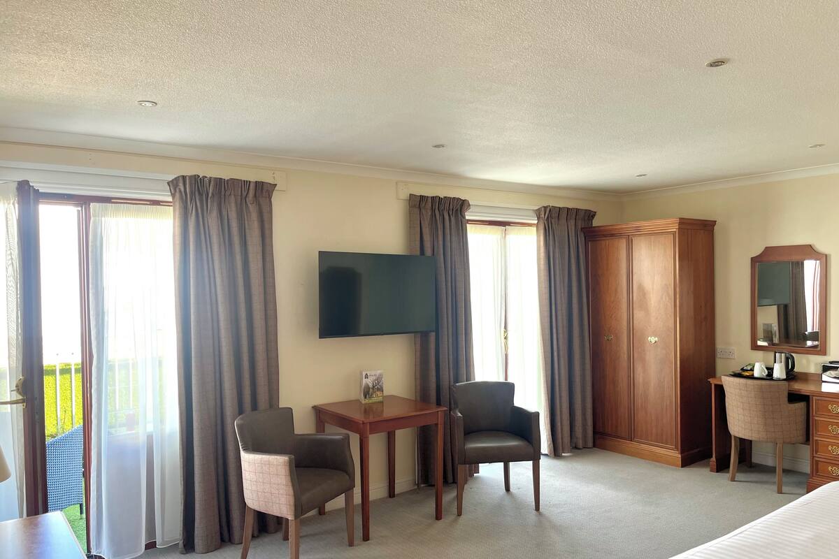 Quarto de hotel com cama, escrivaninha com cadeira, televisão, guarda-roupa e uma janela grande com cortinas.