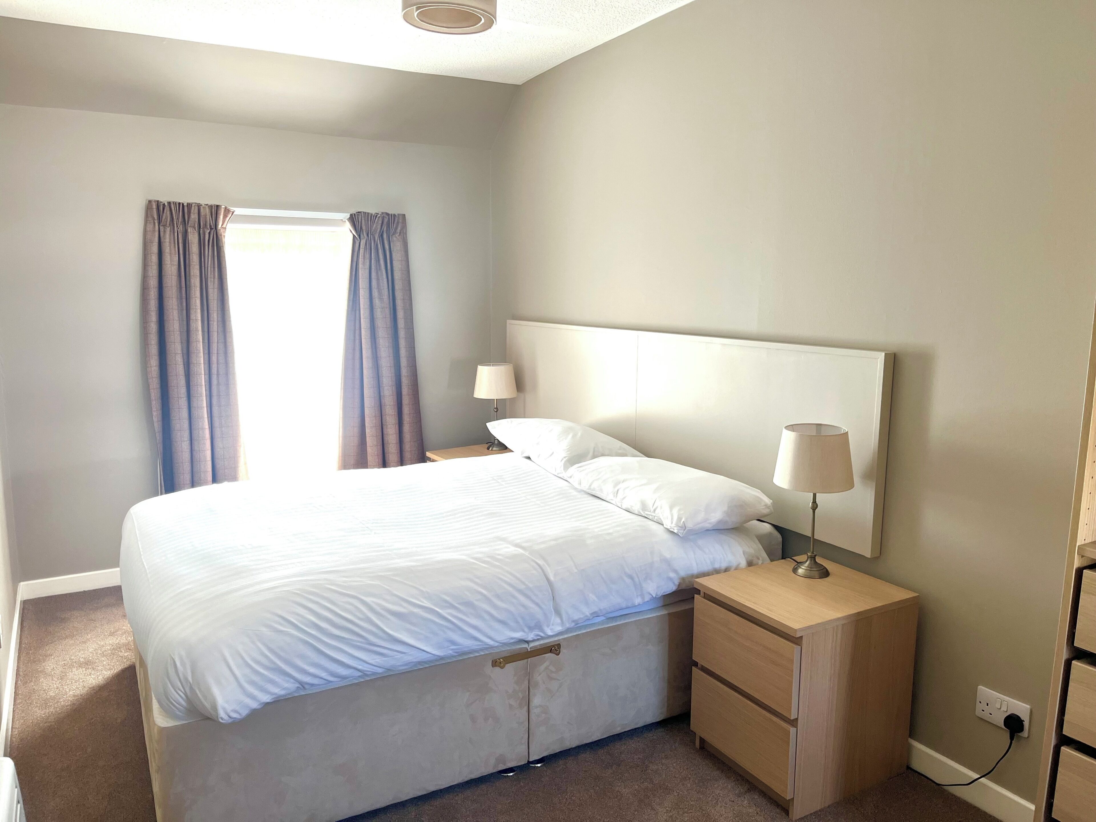 A bedroom with a bed, two bedside tables with lamps, a window with curtains, and a wall socket.
