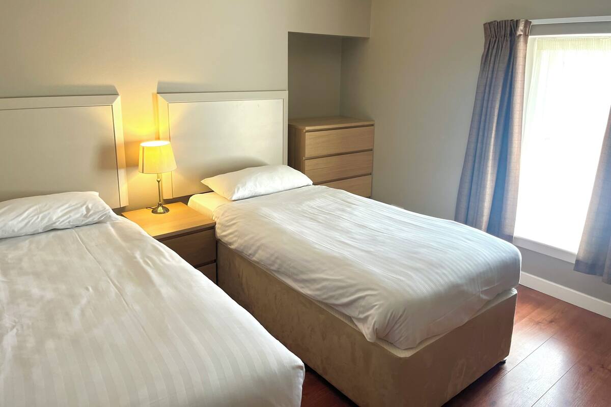Quarto de hotel com duas camas, mesa de cabeceira com abajur, cômoda de madeira e janela com cortinas.