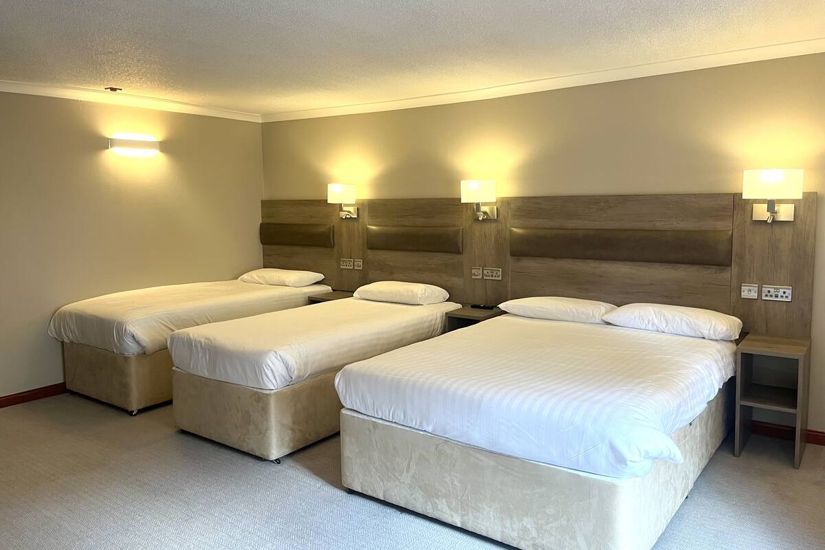 Quarto de hotel com quatro camas de solteiro, cabeceira de madeira, luminárias fixadas na parede e piso coberto por carpete.