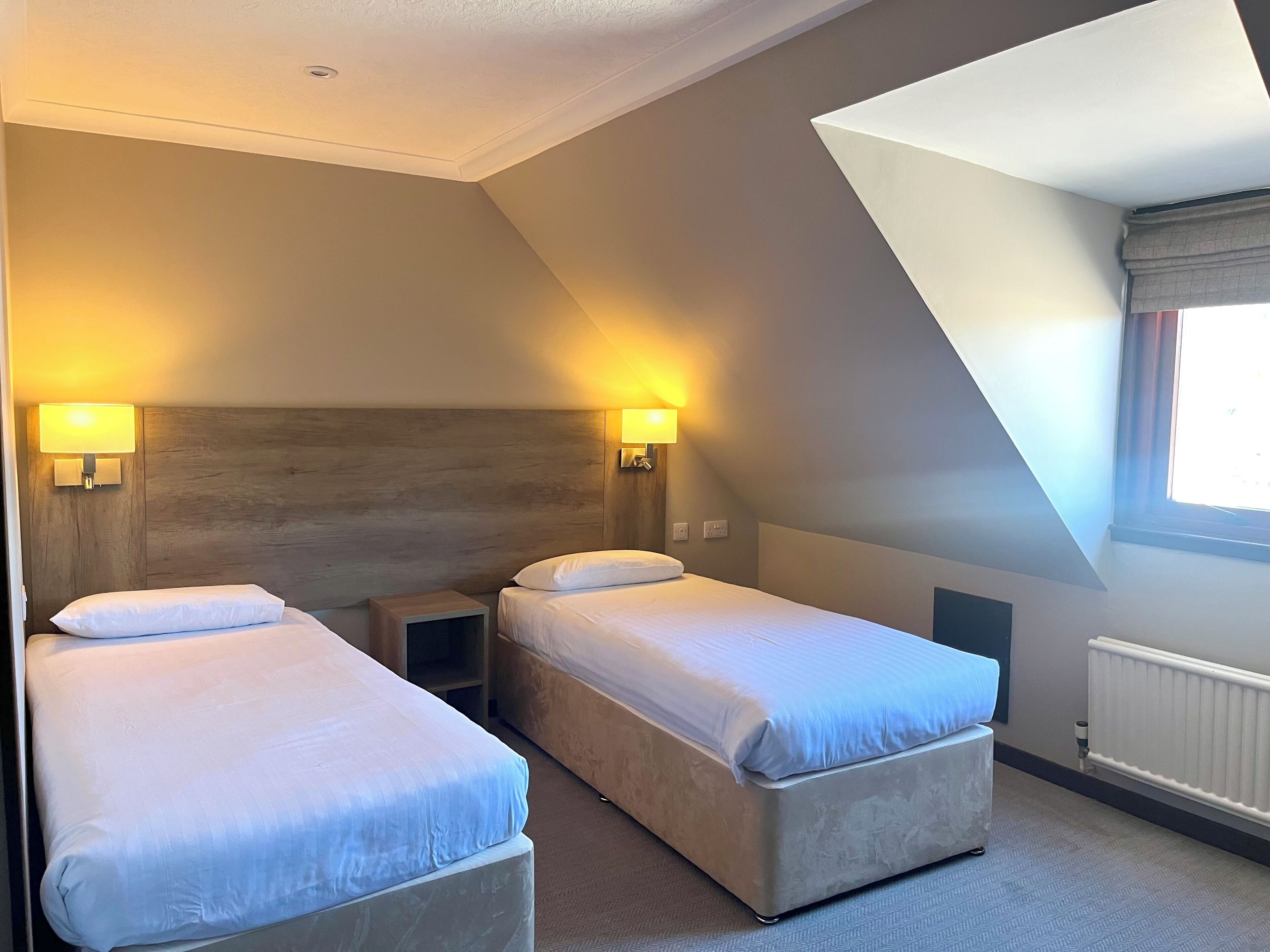 A hotel room with two beds, a wooden headboard, wall-mounted lamps, a radiator, and a sloped ceiling.