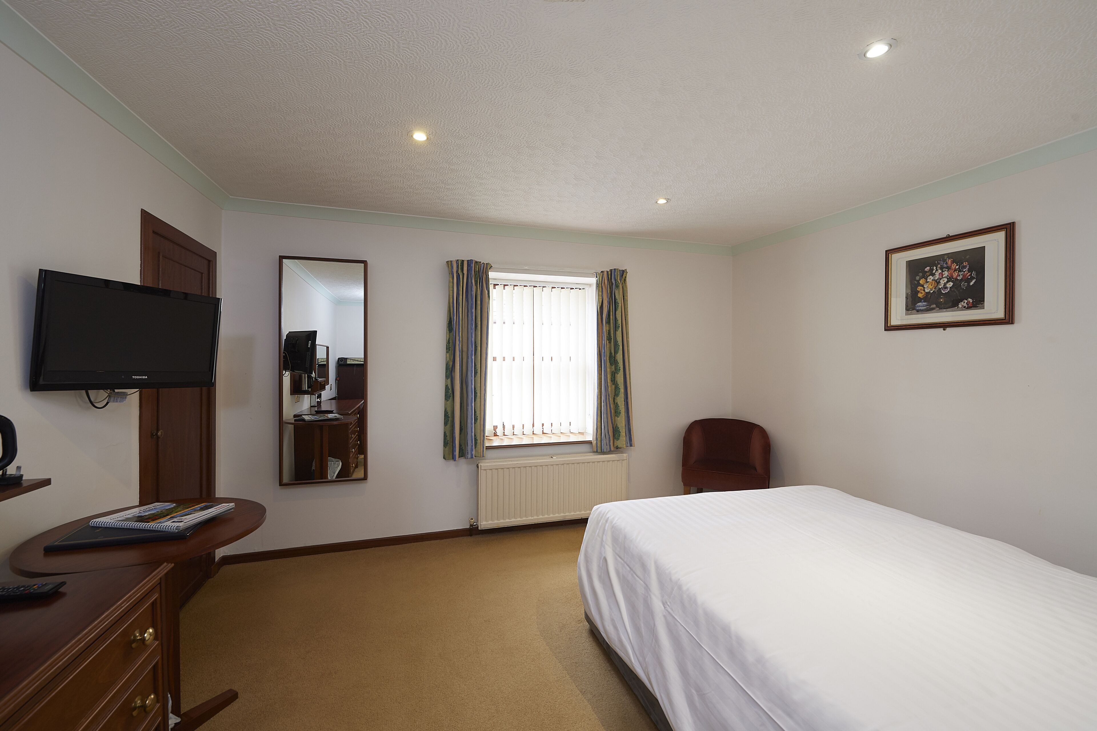 A hotel room with a bed, a desk with a computer, a television, a window with curtains, and a framed picture on the wall.