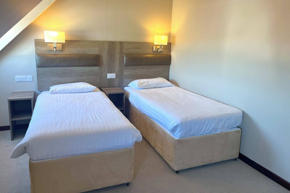 Quarto de hotel com duas camas, cabeceira de madeira, abajures e um criado-mudo pequeno.