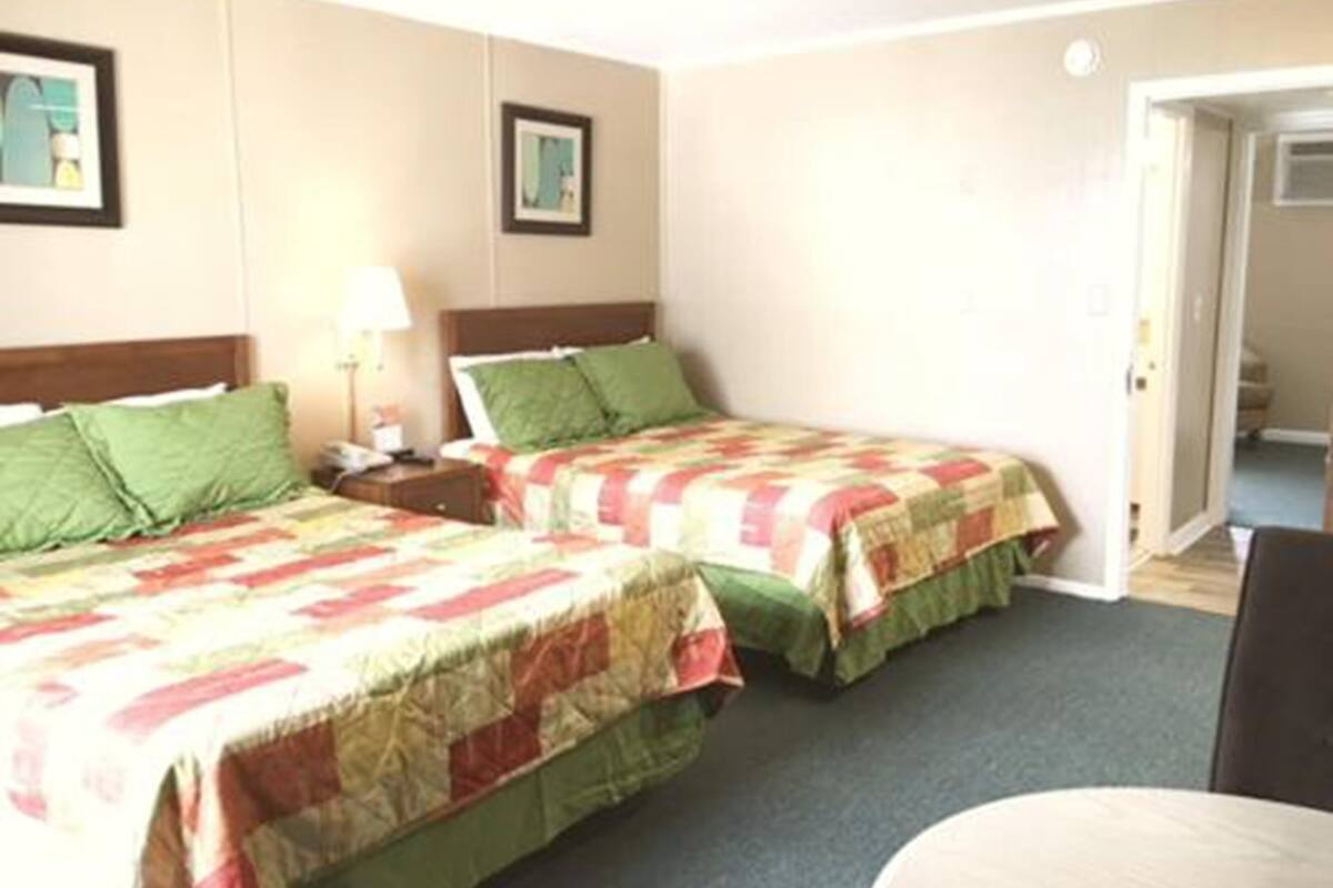 A hotel room with two beds, each with green and red bedding, a bedside table with a lamp, and a door leading to another room.