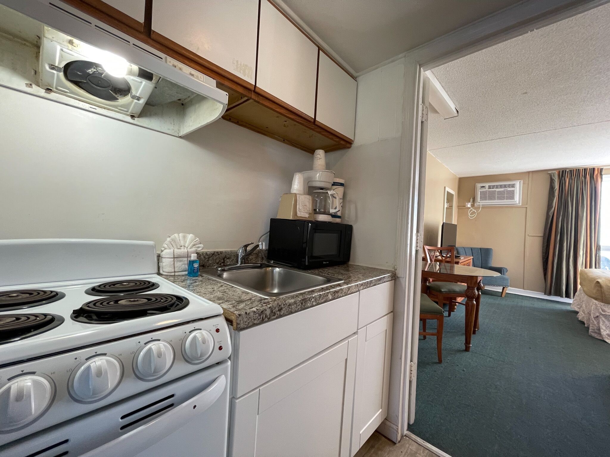A compact kitchen with a stove, sink, and microwave. There is a dining area with a table and chairs visible through an open doorway.
