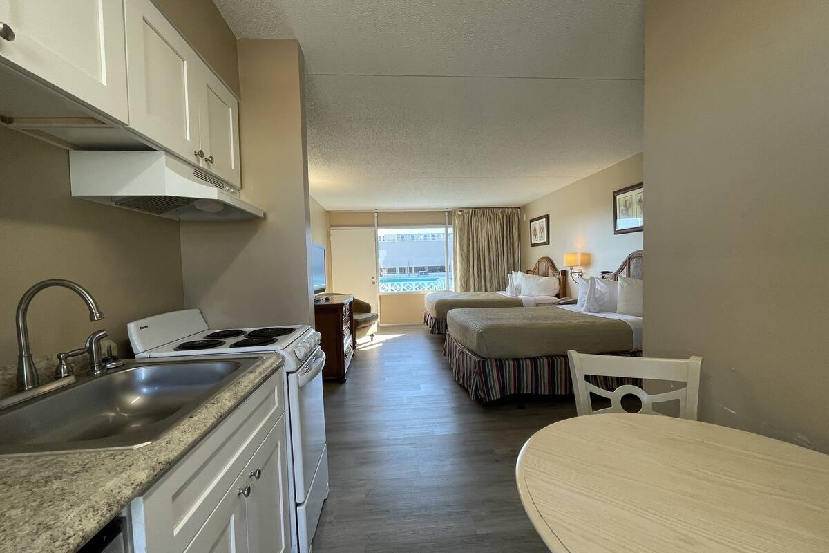 A compact hotel room with a kitchenette, two beds, a small dining table, and a view of the balcony.