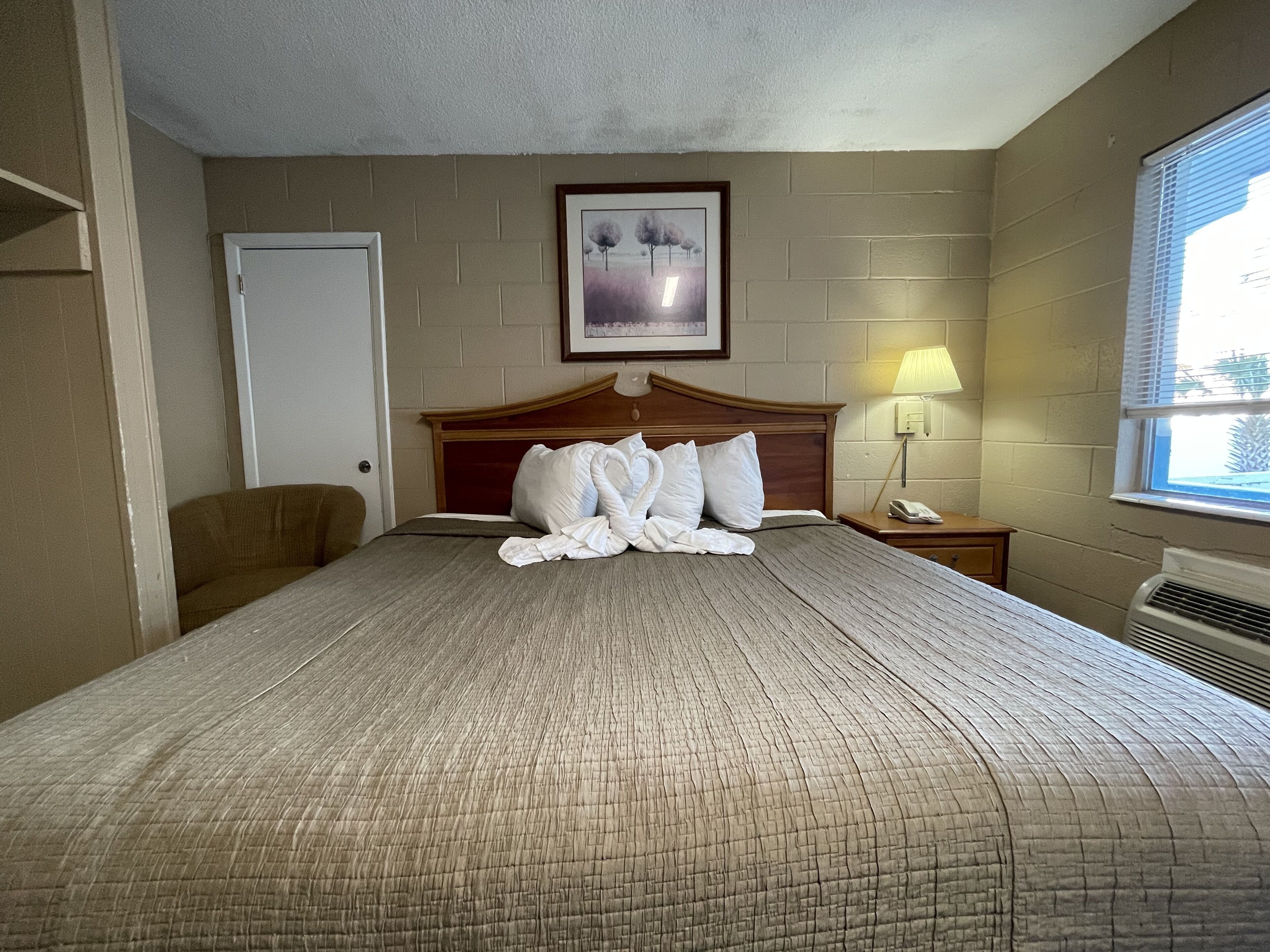 Une chambre d’hôtel avec un grand lit, une tête de lit en bois, un tableau encadré au mur, une lampe de chevet et une fenêtre avec des stores.