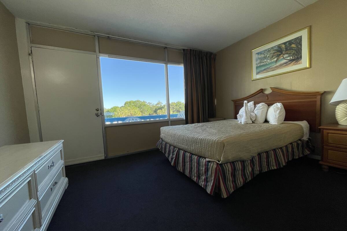A hotel room with a bed, a dresser, a nightstand, a lamp, a painting, and a window with a view of trees and a building.