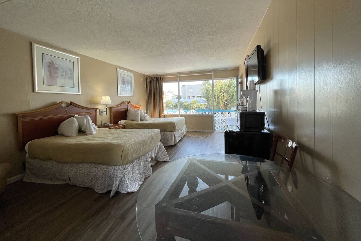 A hotel room with two beds, a glass table, a TV, and a balcony with a view.