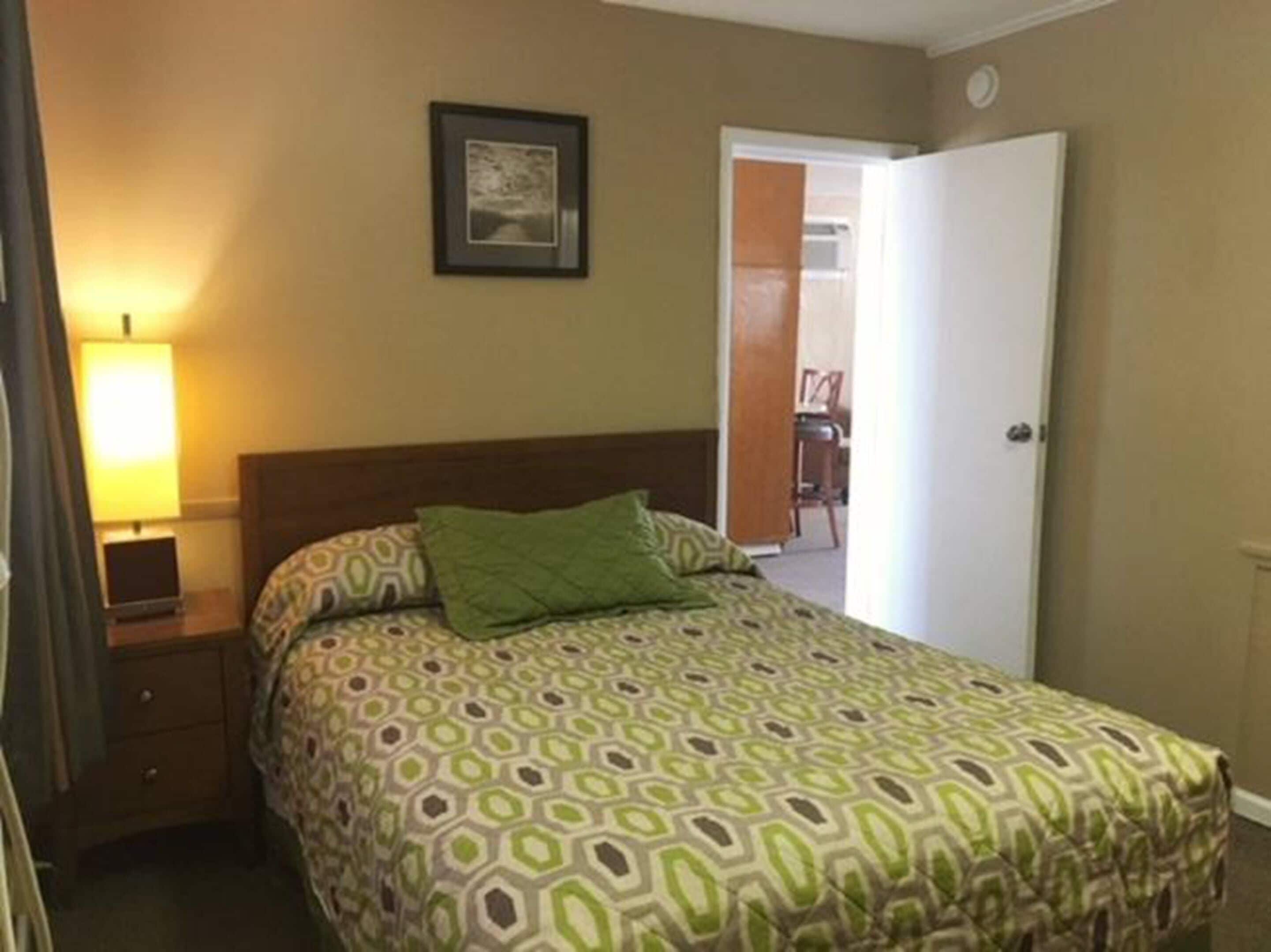 Habitación de hotel con cama, mesita de noche, lámpara, puerta y un cuadro en la pared.