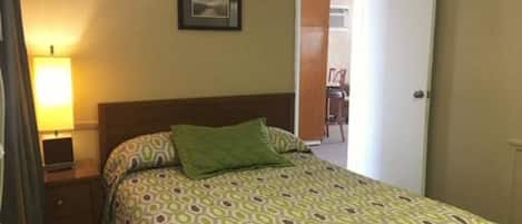 Habitación de hotel con cama, mesita de noche, lámpara, puerta y un cuadro en la pared.