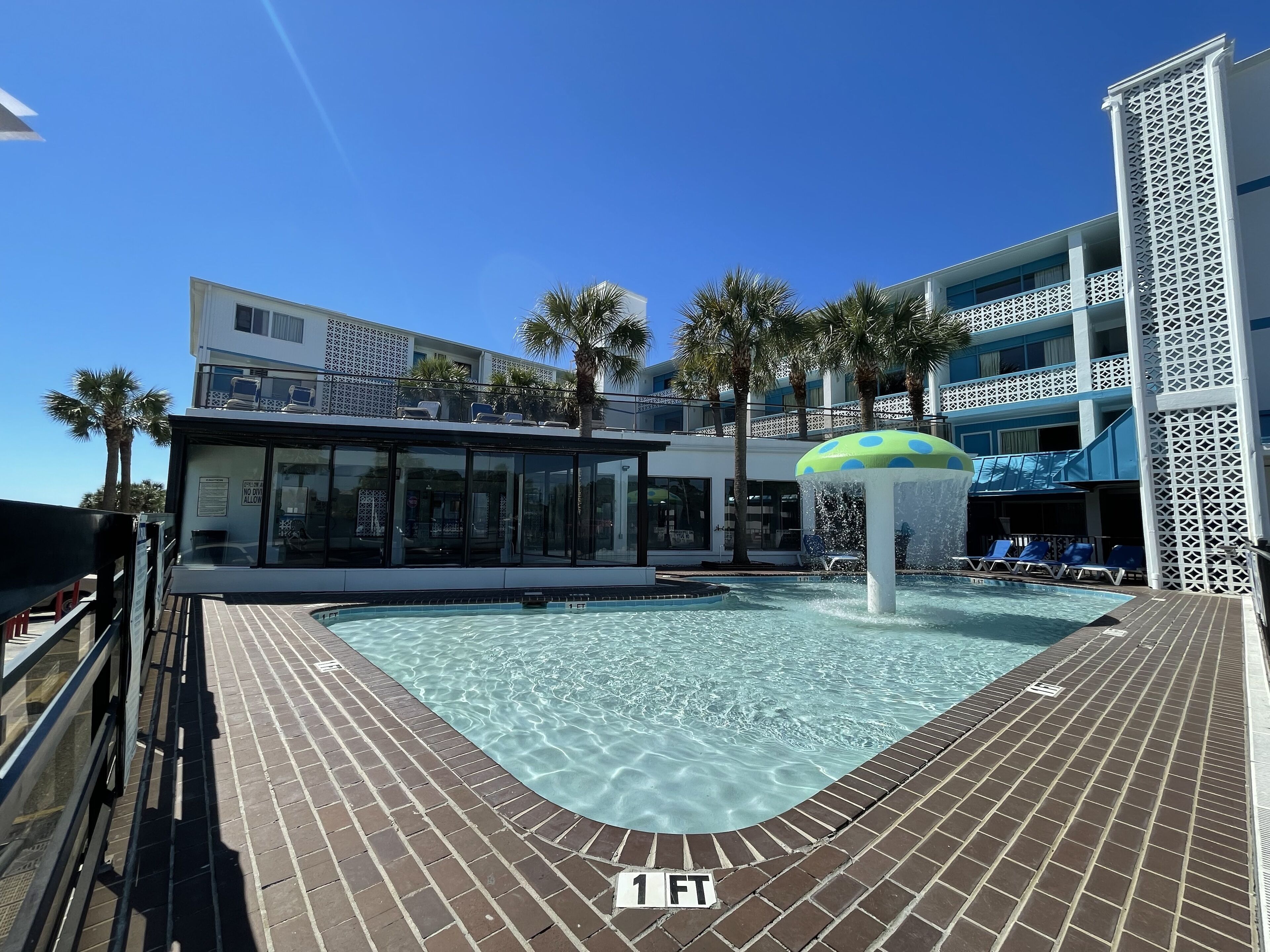 A pool area with a clear water surface, a sign indicating a depth of 1 FT, and a building with a balcony and palm trees in the background.