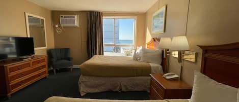A hotel room with two beds, a television, a chair, a desk with a lamp, and a window with a view of the beach.
