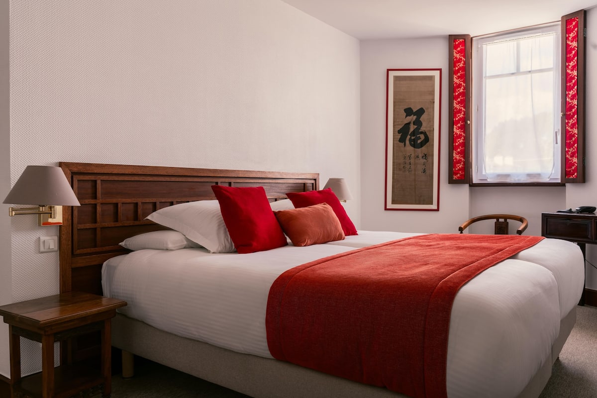 A neatly made bed with red and white bedding, a wooden headboard, and a red throw blanket. There is a bedside table with a lamp, a framed artwork on the wall, and a window with red curtains.