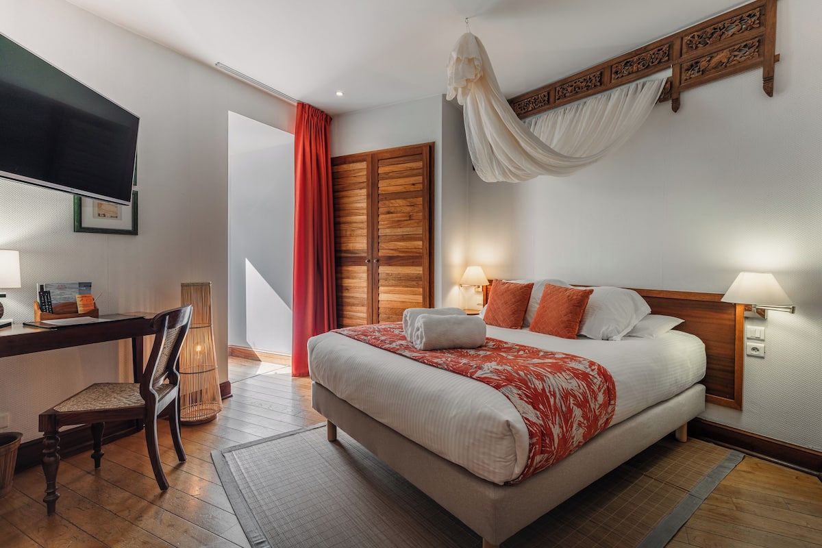 A hotel room with a bed, a desk with a chair, a TV, and a large window with curtains.