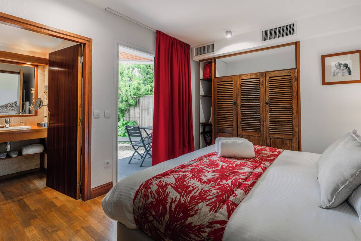A bedroom with a bed, a wooden wardrobe, a red curtain, and a bathroom with a mirror and sink.