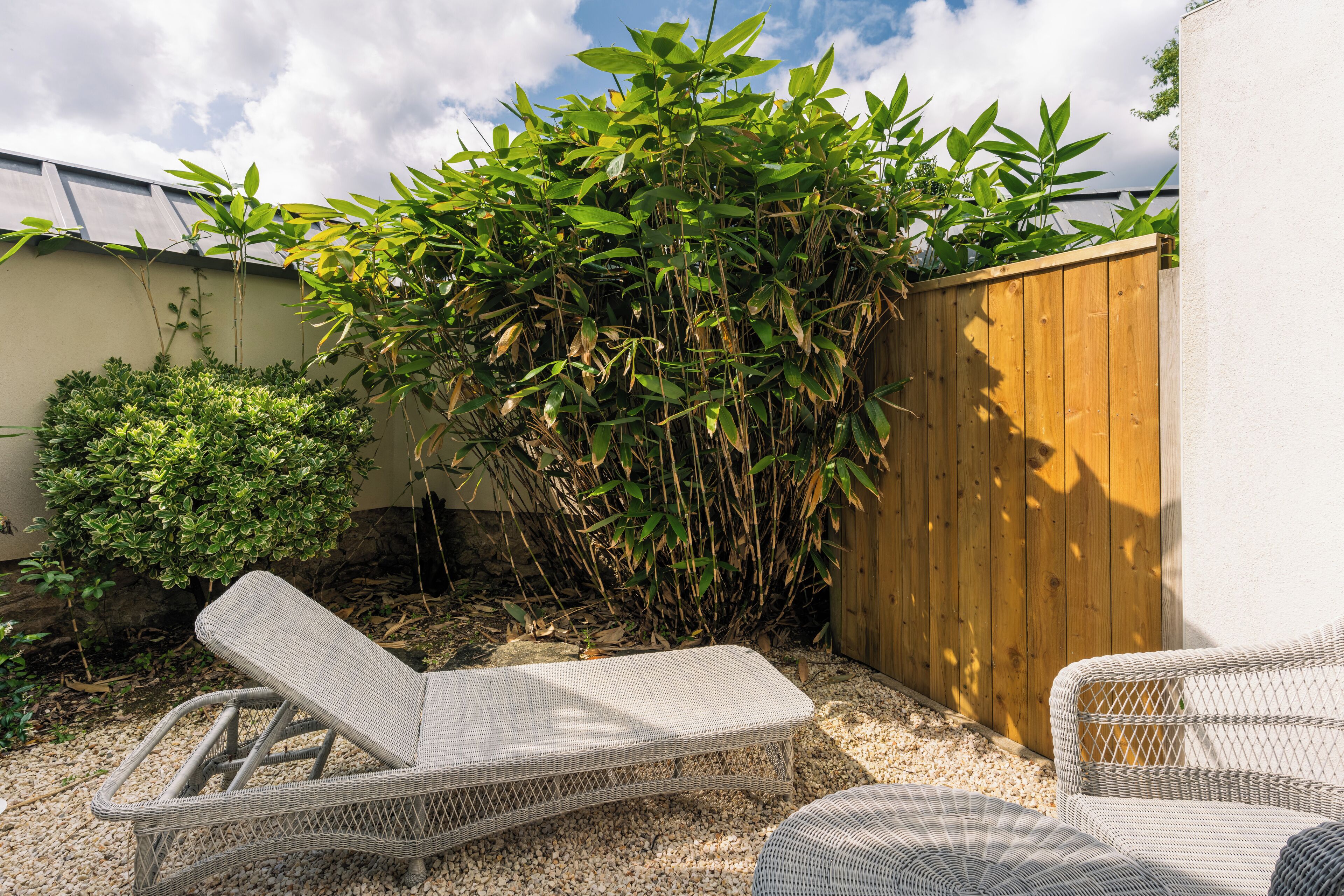 A patio area with wicker lounge chairs, a wooden fence, and lush greenery.