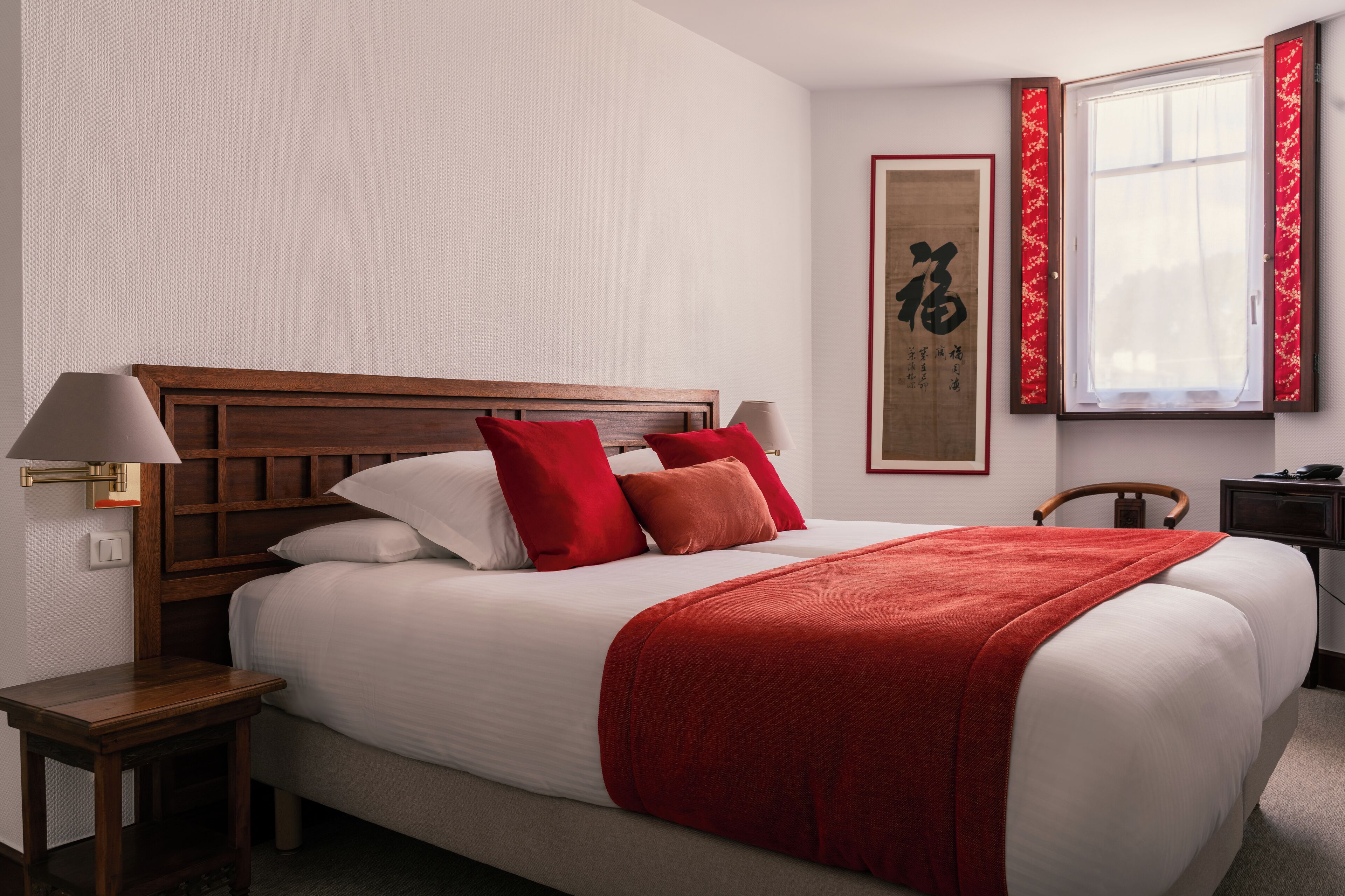 A neatly made bed with a red blanket and pillows, a wooden headboard, a bedside table with a lamp, and a framed artwork on the wall.
