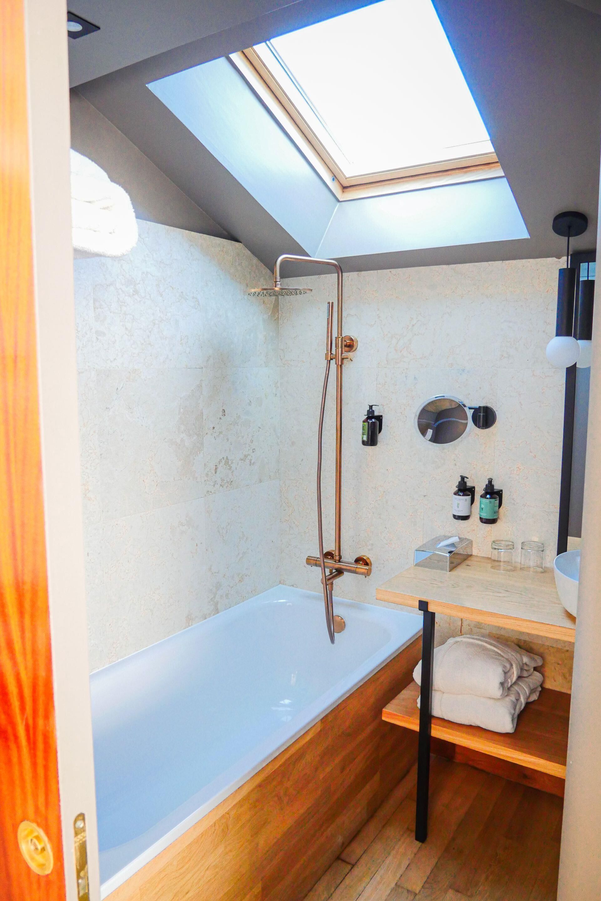 Un baño pequeño con tragaluz, una bañera, un estante de madera con toallas y un espejo.