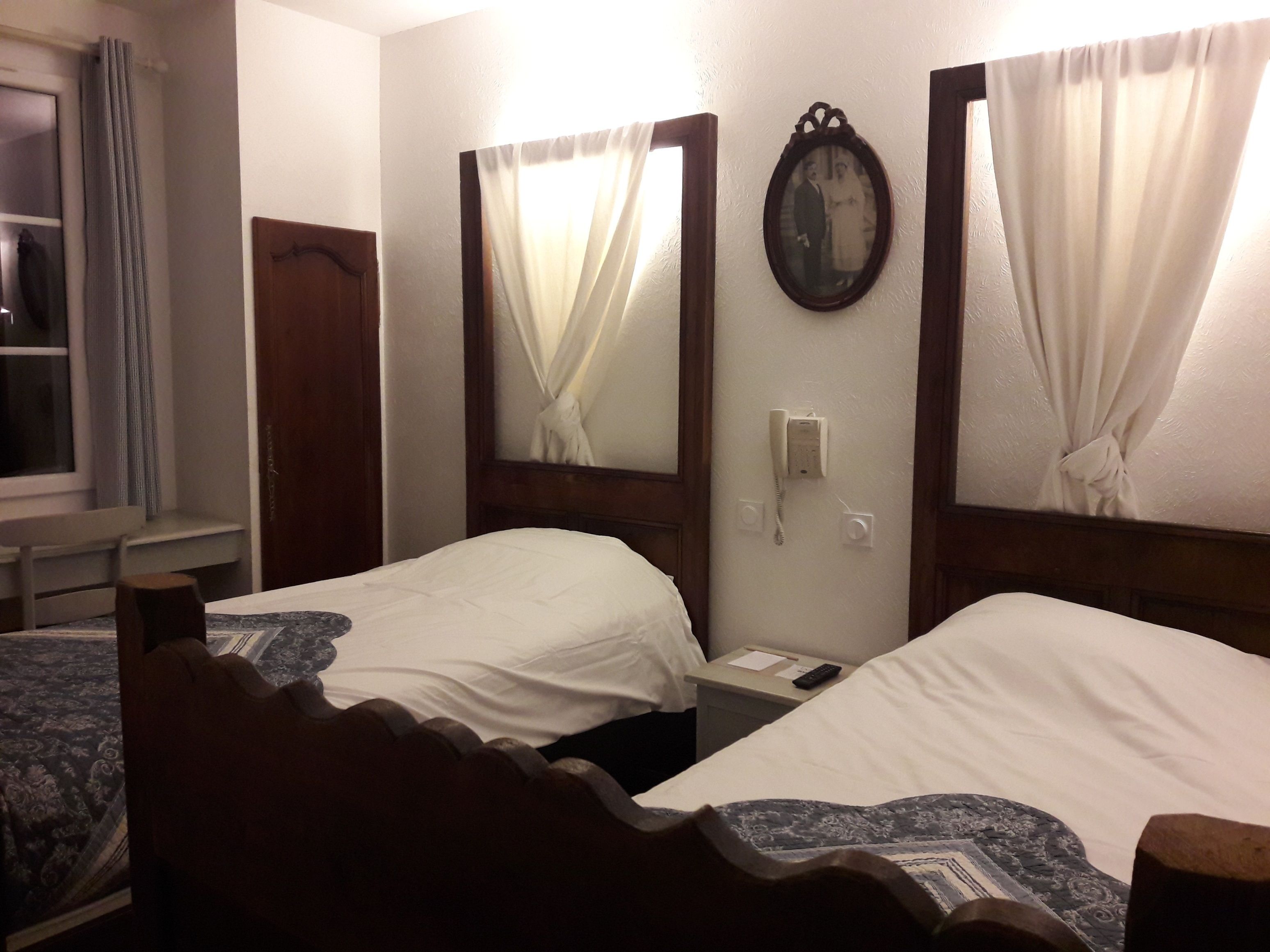 A hotel room with two single beds, a wooden headboard, a bedside table with a phone and remote control, and a window with curtains.