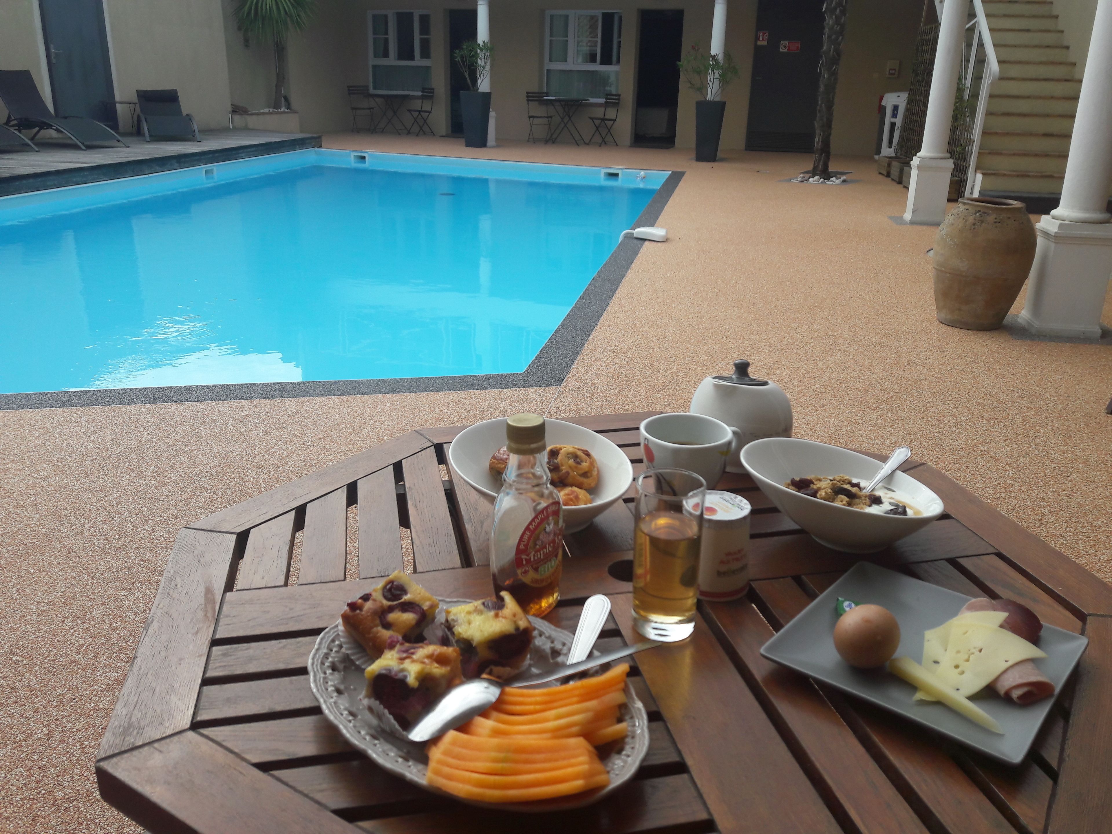 A poolside dining area with a table set for breakfast, including pastries, fruit, and beverages.