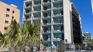 Un hôtel à plusieurs étages doté de balcons, avec un palmier au premier plan et un ciel bleu dégagé.