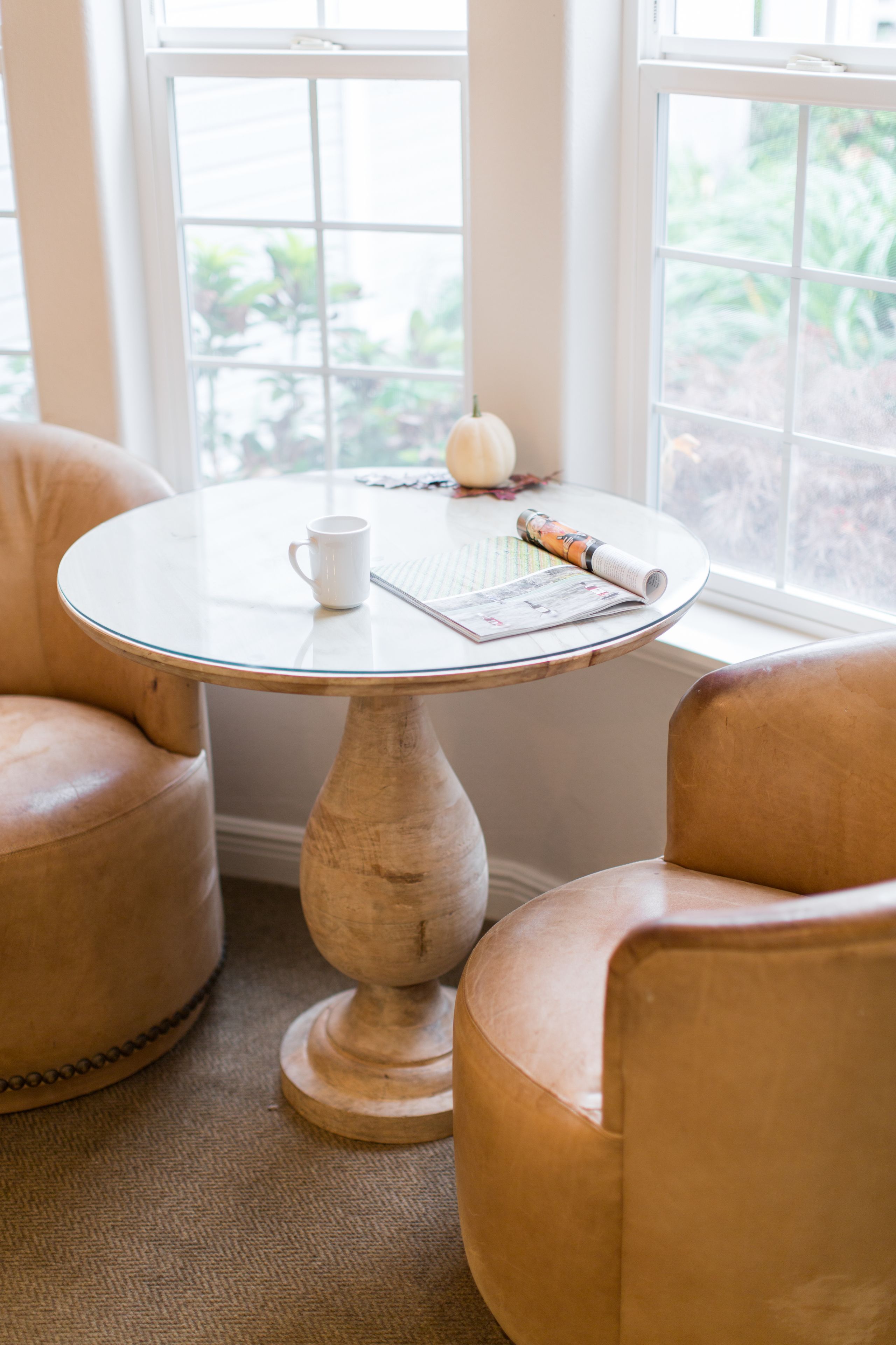 A round wooden table with a white mug and a rolled newspaper, surrounded by two leather armchairs, with a window in the background.