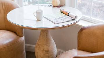 A round wooden table with a white mug and a rolled newspaper, surrounded by two leather armchairs, with a window in the background.