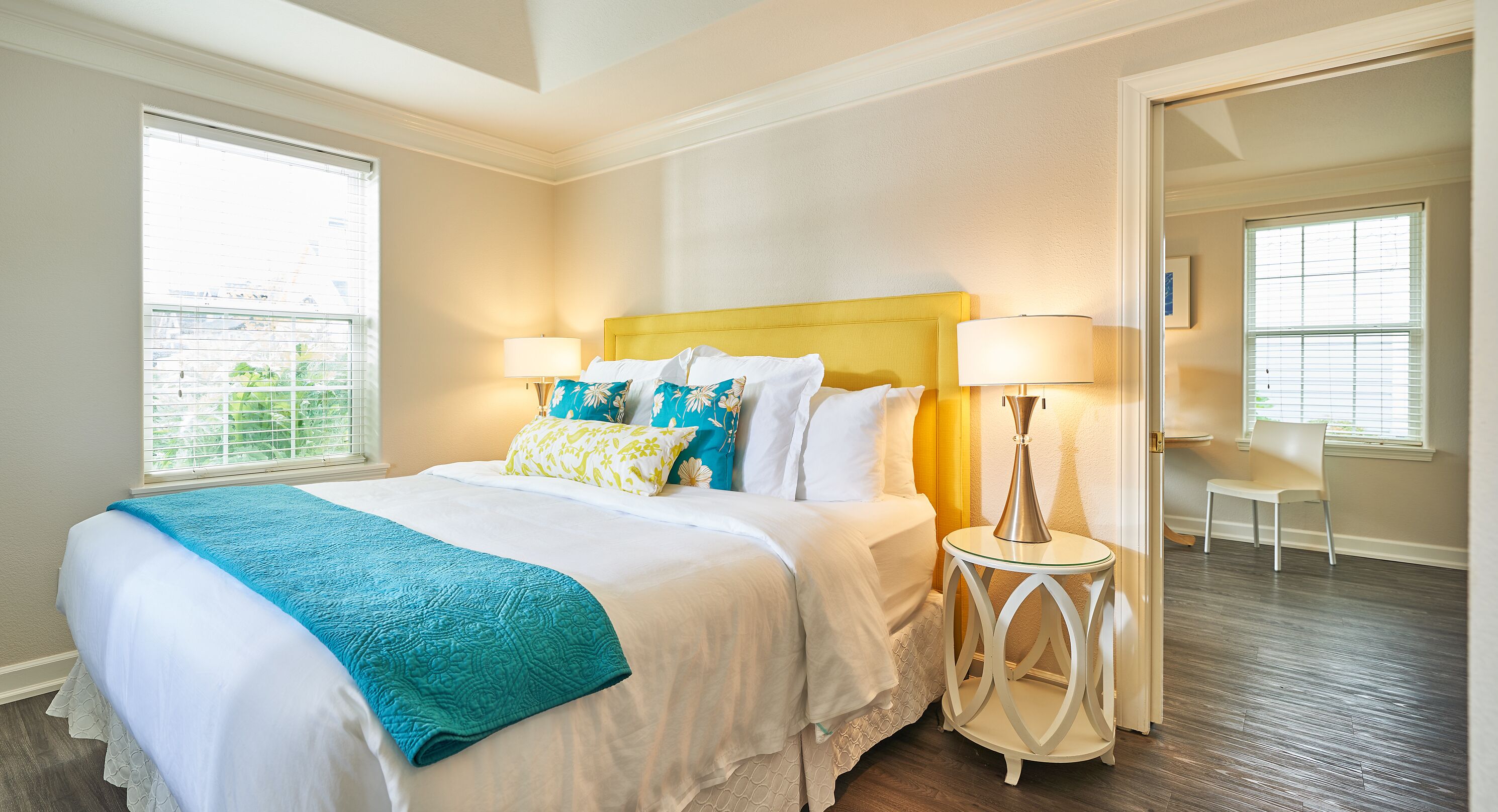 A hotel room with a large bed, two bedside lamps, a view of the outdoors, and a door leading to another room.