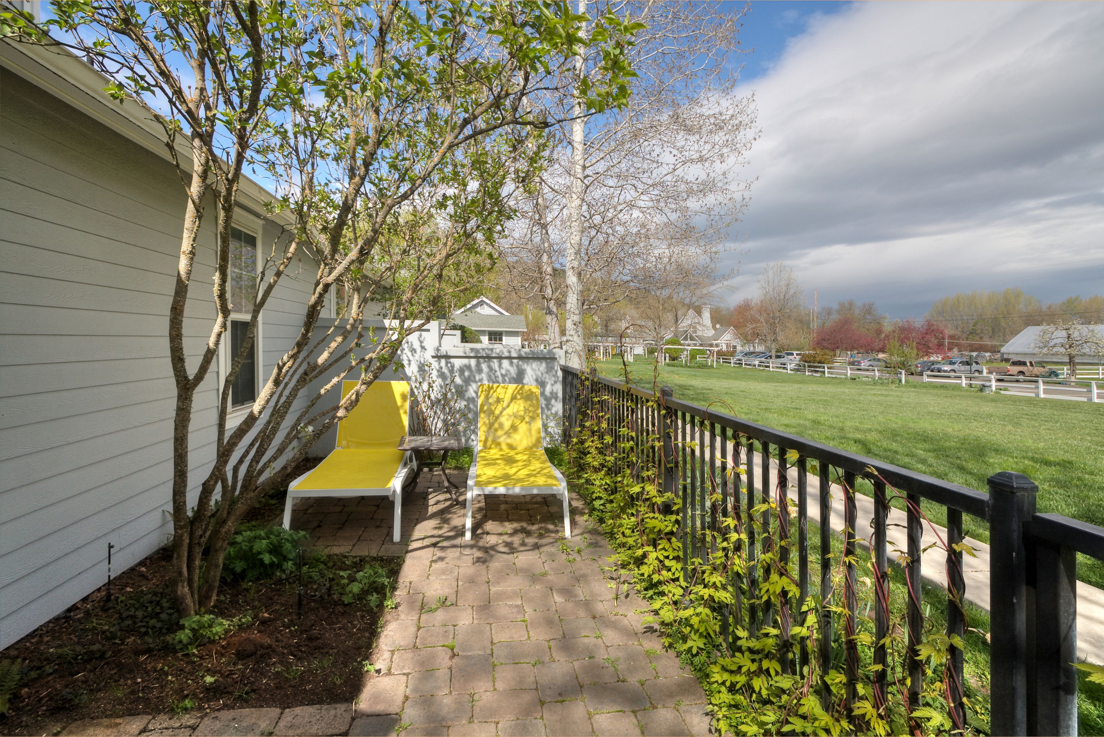 A paved patio with a yellow chair and a white chair, a small tree, and a view of a green field and buildings in the distance.