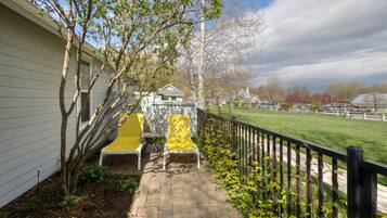 A paved patio with a yellow chair and a white chair, a small tree, and a view of a green field and buildings in the distance.