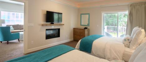A bedroom with two beds, a TV mounted on the wall, a window with a view of greenery, and a ceiling fan.