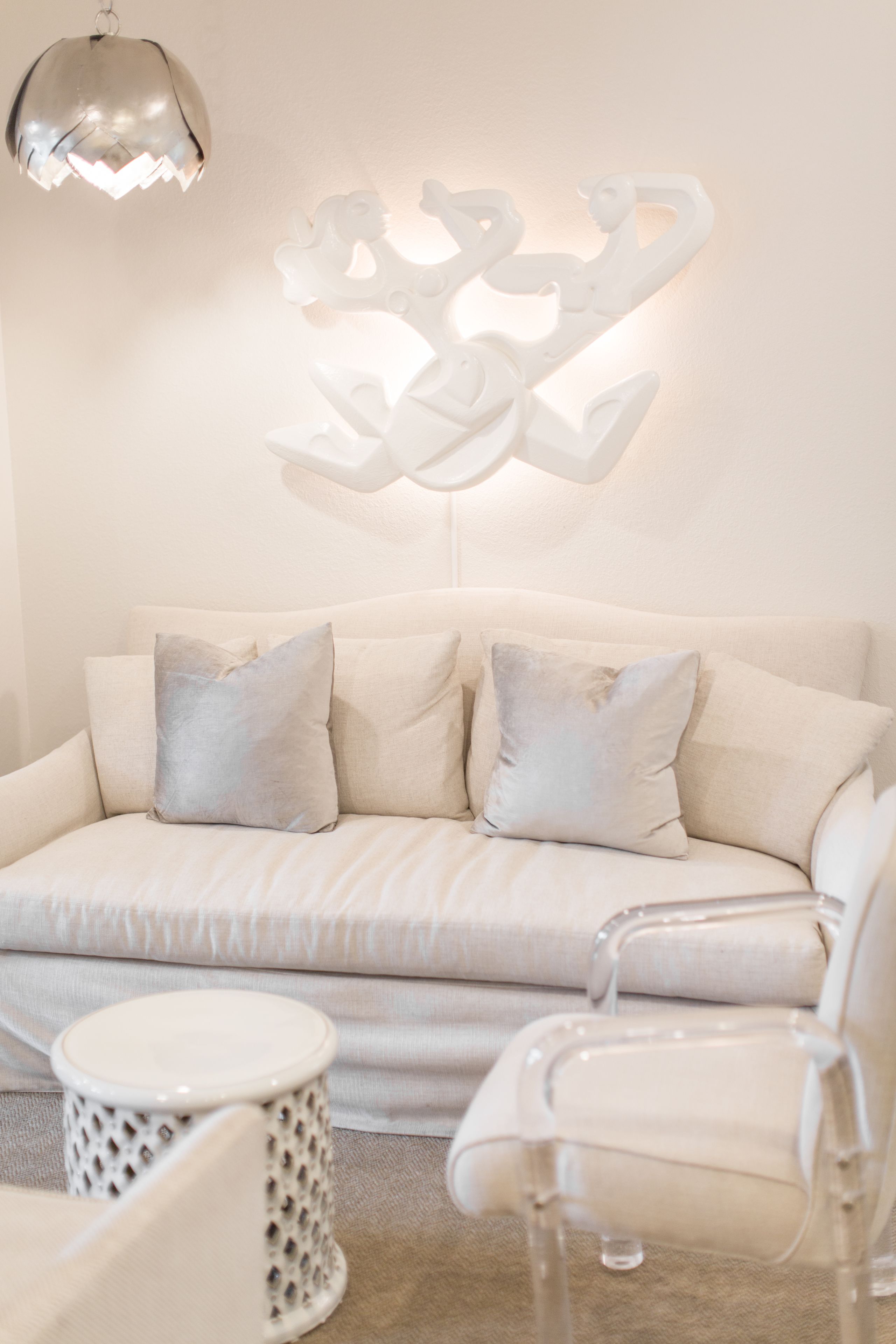 A modern living room with a white sofa, two clear chairs, a small round table, and a decorative wall piece.