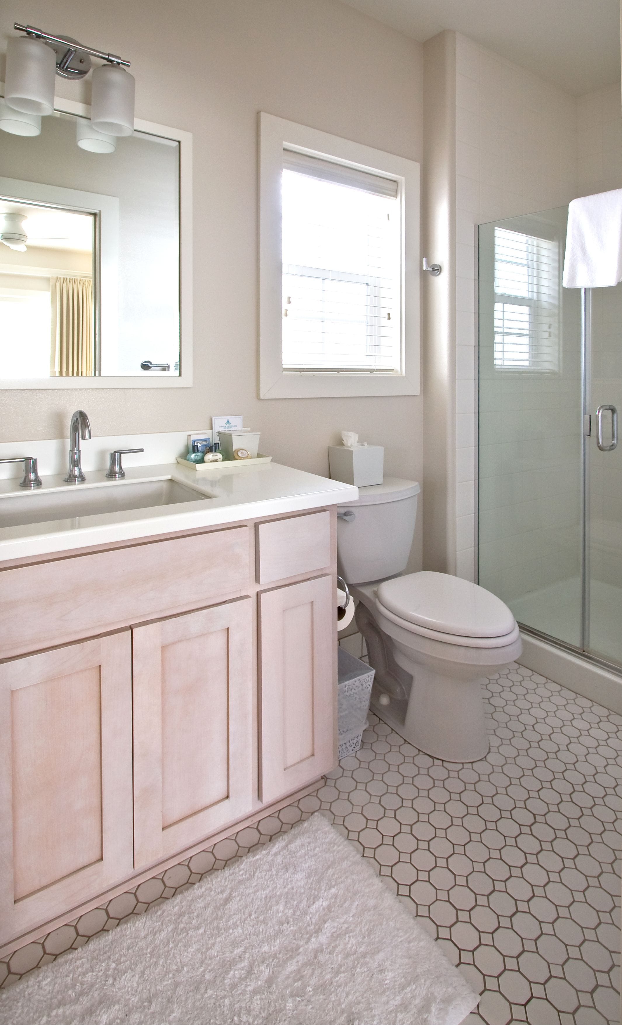 Un baño con lavamanos blanco, espejo y retrete, cabina de ducha de vidrio y baldosas hexagonales.