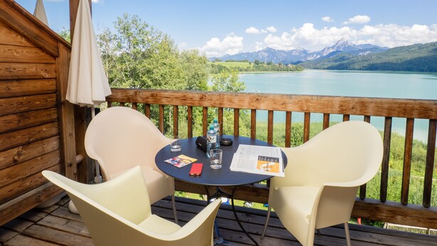 A wooden deck with two chairs and a table, overlooking a lake with mountains in the background. - Appartementhotel Seespitz (Fuessen)