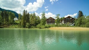 A lakeside hotel with multiple floors, surrounded by trees and a clear sky. - Appartementhotel Seespitz (Fuessen)