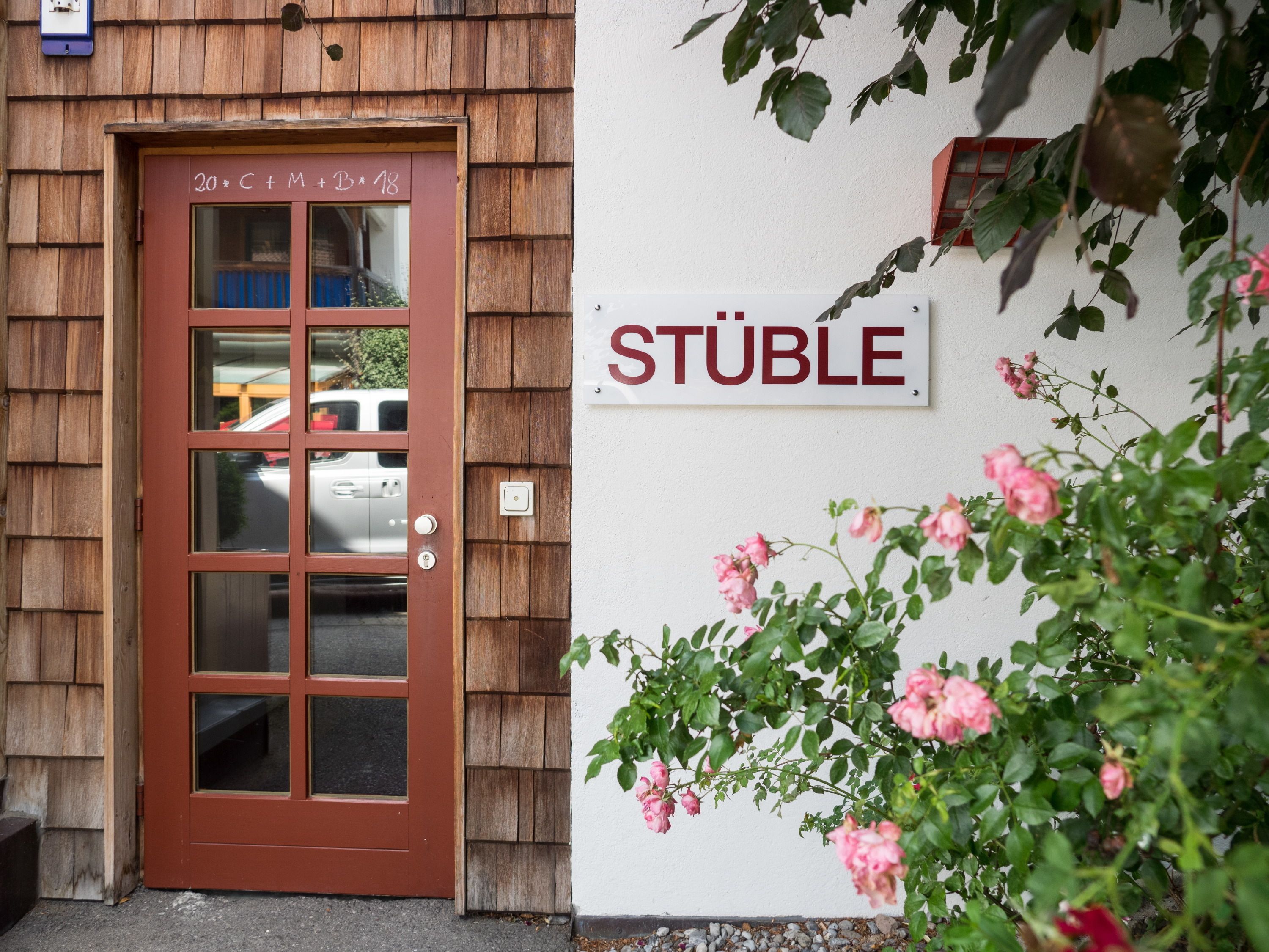 A wooden door with a window, a sign reading "STÜBLE", and pink roses in the foreground.