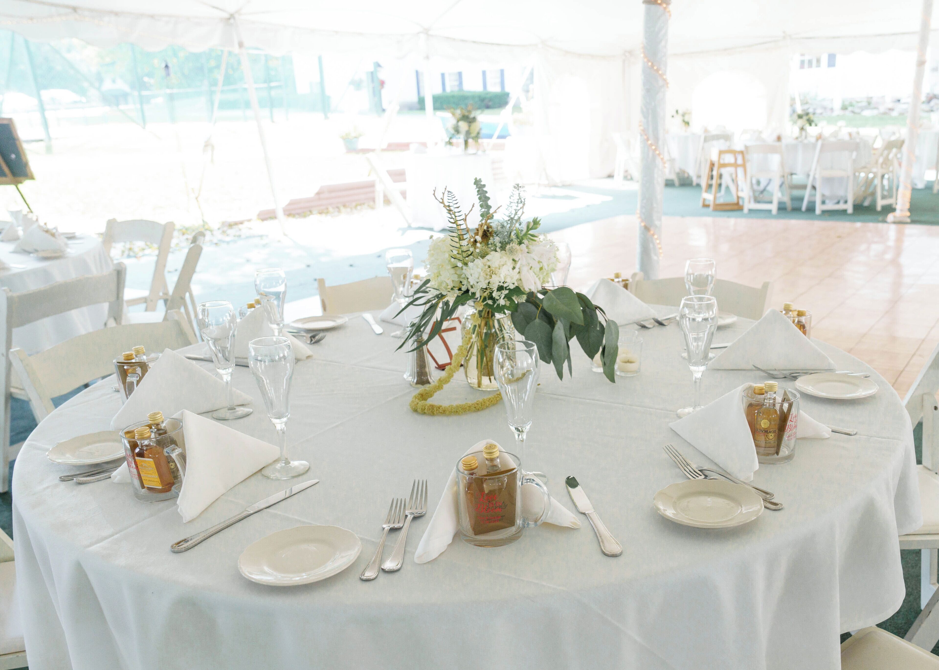 Una mesa redonda dispuesta para un evento formal, con manteles blancos, cristalería y un centro de mesa con flores.