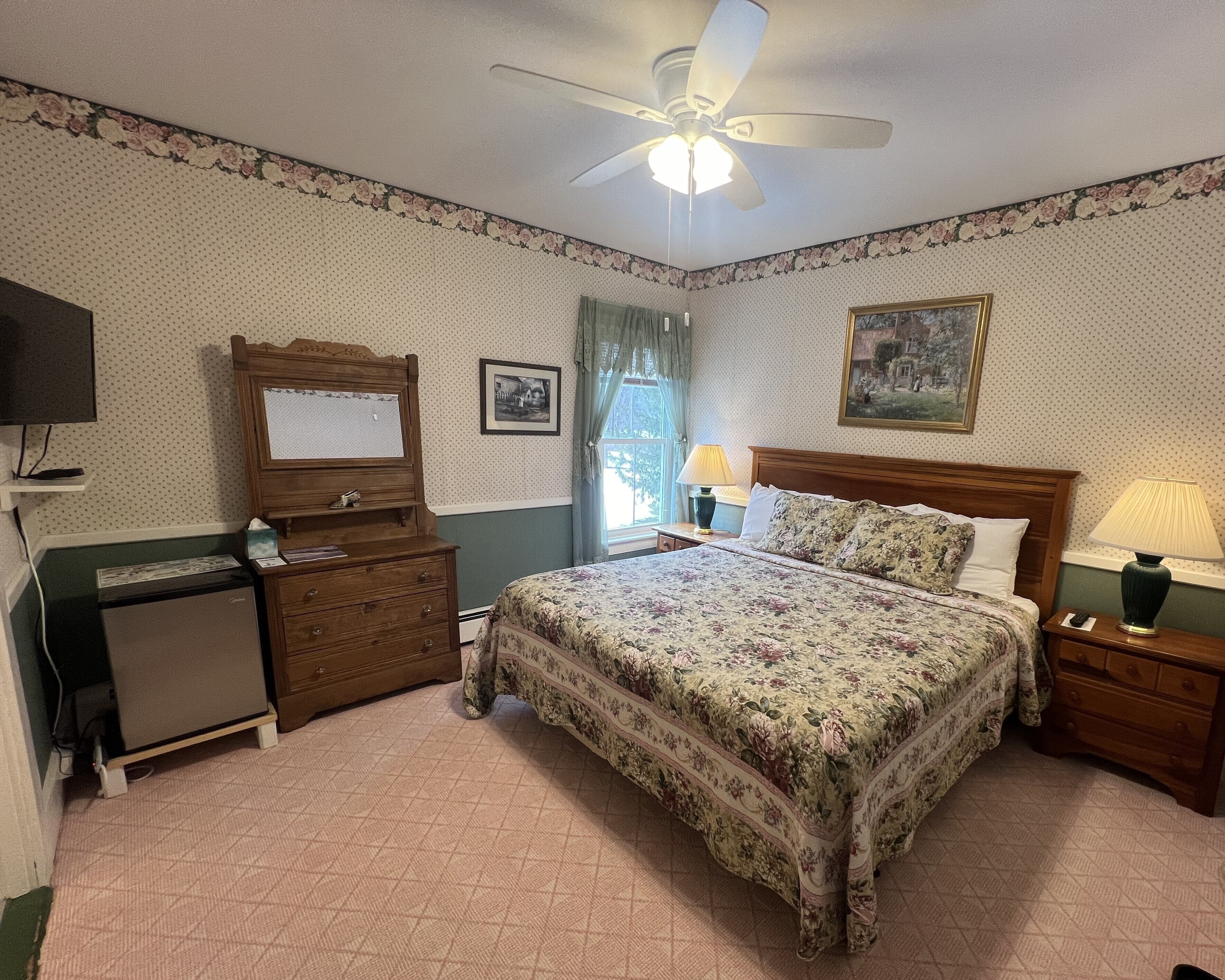 Une chambre à coucher comprenant un lit, un bureau, un ventilateur de plafond, une fenêtre avec des rideaux et un téléviseur fixé au mur.
