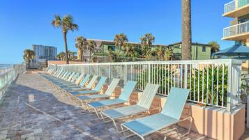 A beachside walkway with lounge chairs, palm trees, and a building in the background.