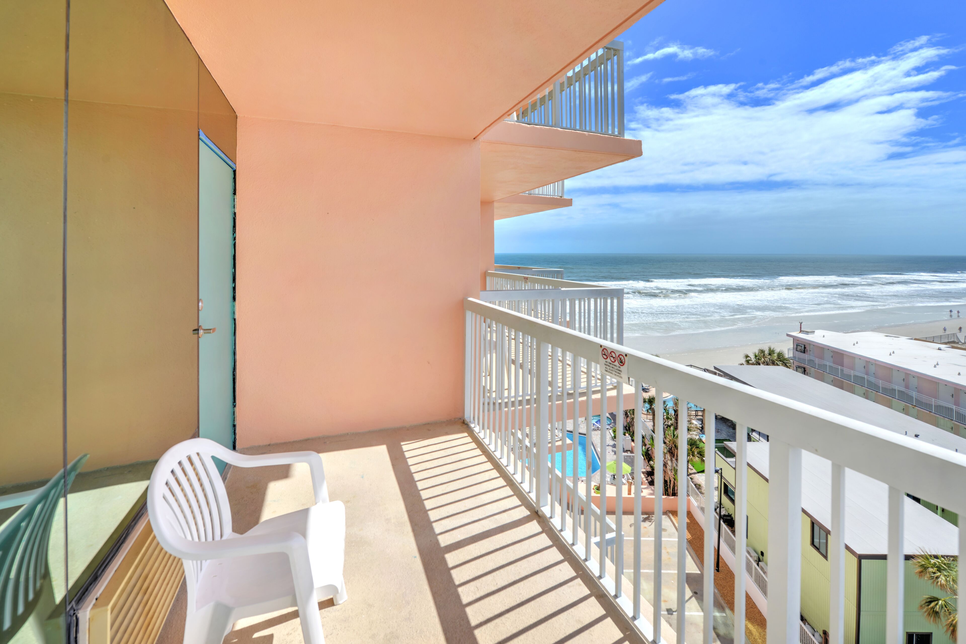 A balcony with a view of the ocean, a pool area, and a white chair.