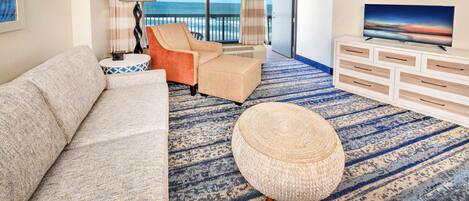 A hotel room with a sofa, armchair, ottoman, a TV on a dresser, and a view of the ocean through large windows.
