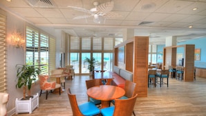 A spacious indoor area with a round wooden table surrounded by wicker chairs with blue cushions, a seating area with a bench, and a large window with blinds. - Bahama House (Daytona Beach Shores)